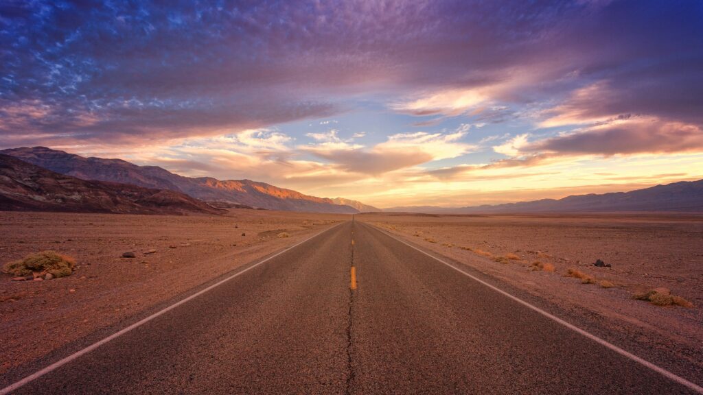 Road leading into the sunset, surrounded by mountains and open sky.